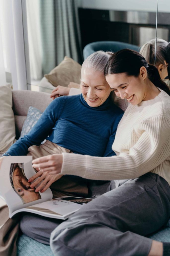 Two women sit closely on a couch, smiling and looking at a magazine together. One points at a page while the other leans in, both appearing engaged and relaxed.