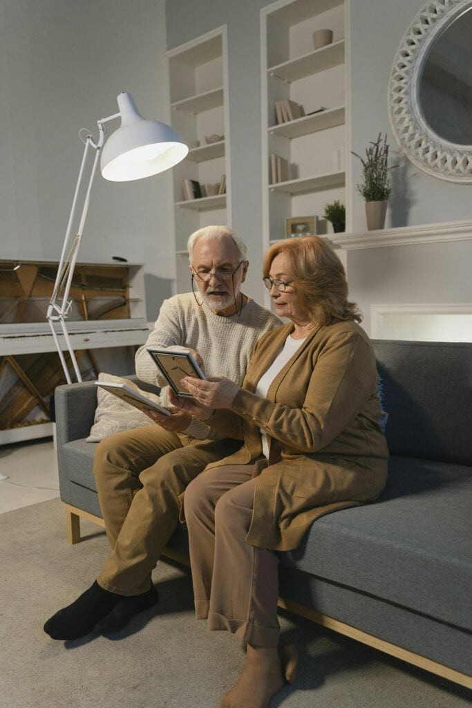An elderly couple sits on a gray sofa in a living room, looking at framed photos together under a floor lamp.