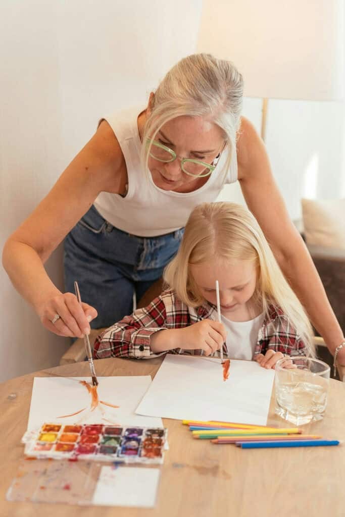 An older woman helps a young girl paint with watercolors at a table. Both are focused on their artwork, with painting supplies and a glass of water nearby.