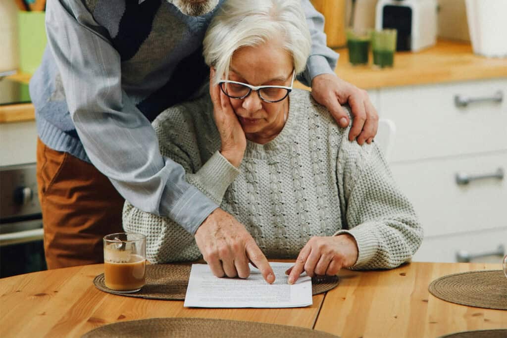 An older woman sits at a table looking at paperwork while an older man stands beside her, pointing at the document.
