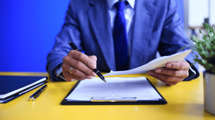 A person in a suit sits at a yellow desk, reviewing and signing documents with a pen, with a notebook, tablet, and small plant nearby.