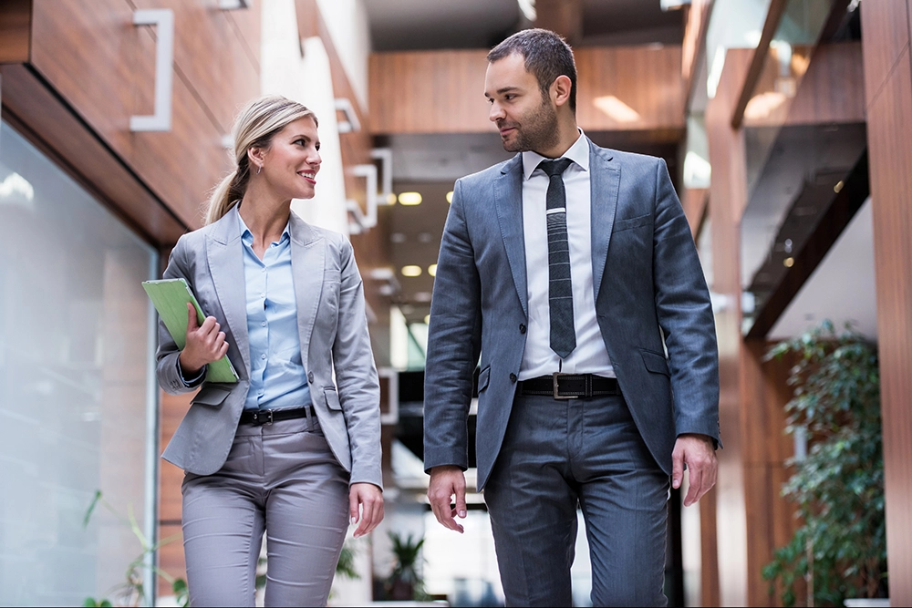 A woman and a man in business attire walk through a modern office hallway, talking and holding documents.