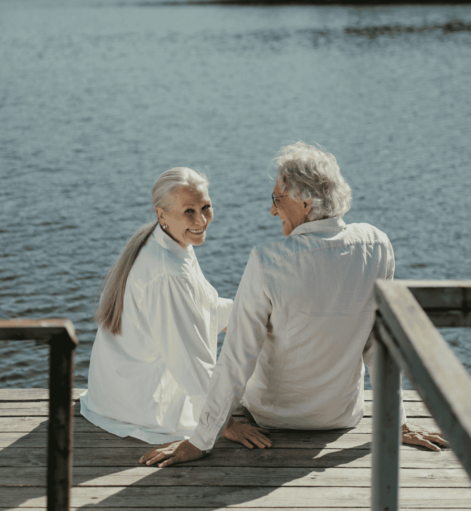 An elderly couple sits on a wooden dock by the water, both dressed in light-colored clothing. The woman is smiling at the camera while the man looks at her.
