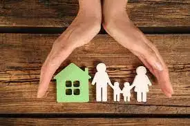 Two hands form a protective gesture over paper cutouts of a house and a family of four, placed on a wooden surface.
