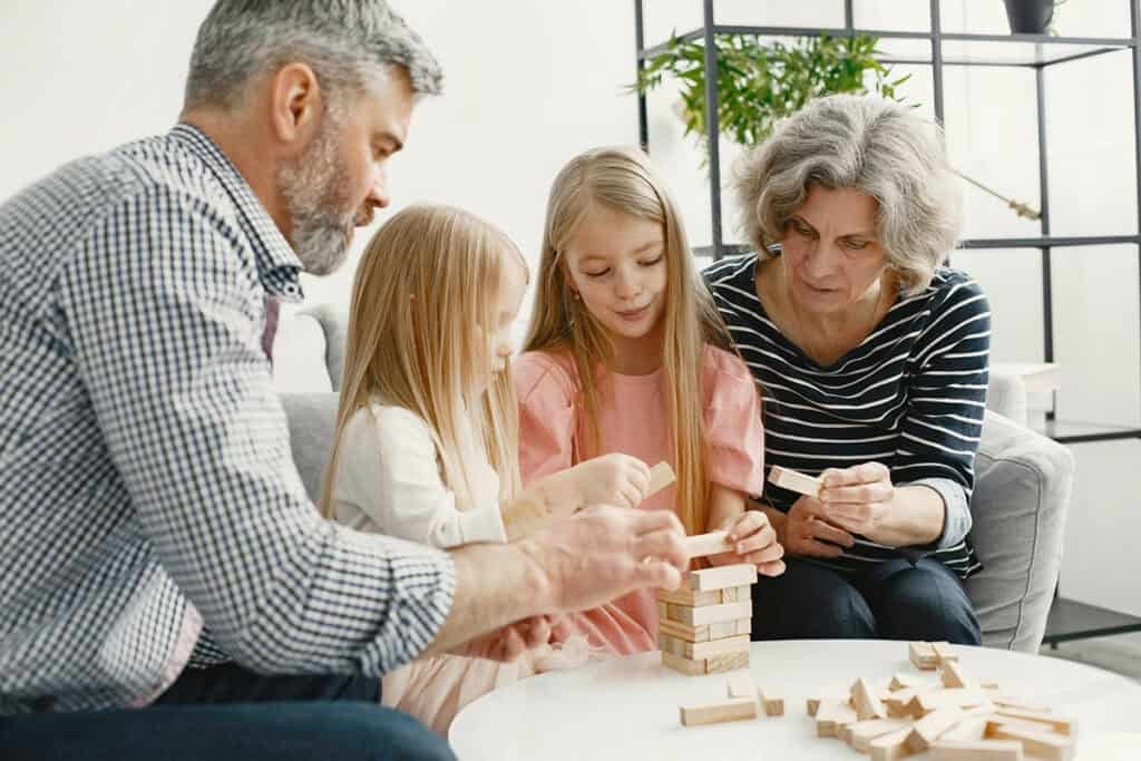 Two adults and two children sit around a table playing a wooden block stacking game together in a living room.