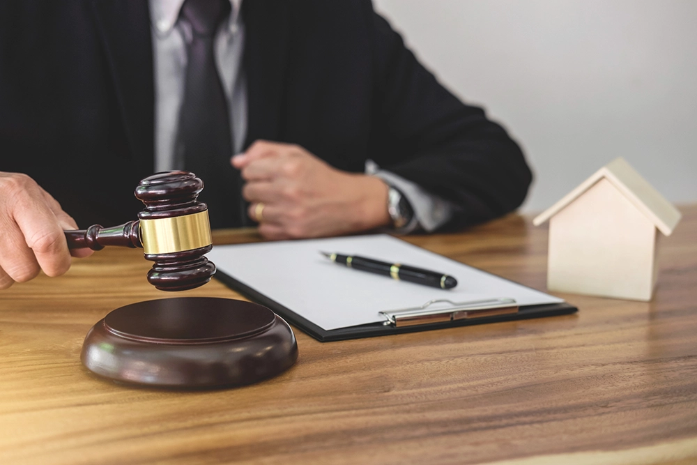 A person in a suit sits at a desk with a gavel, clipboard, pen, and a small wooden house model.