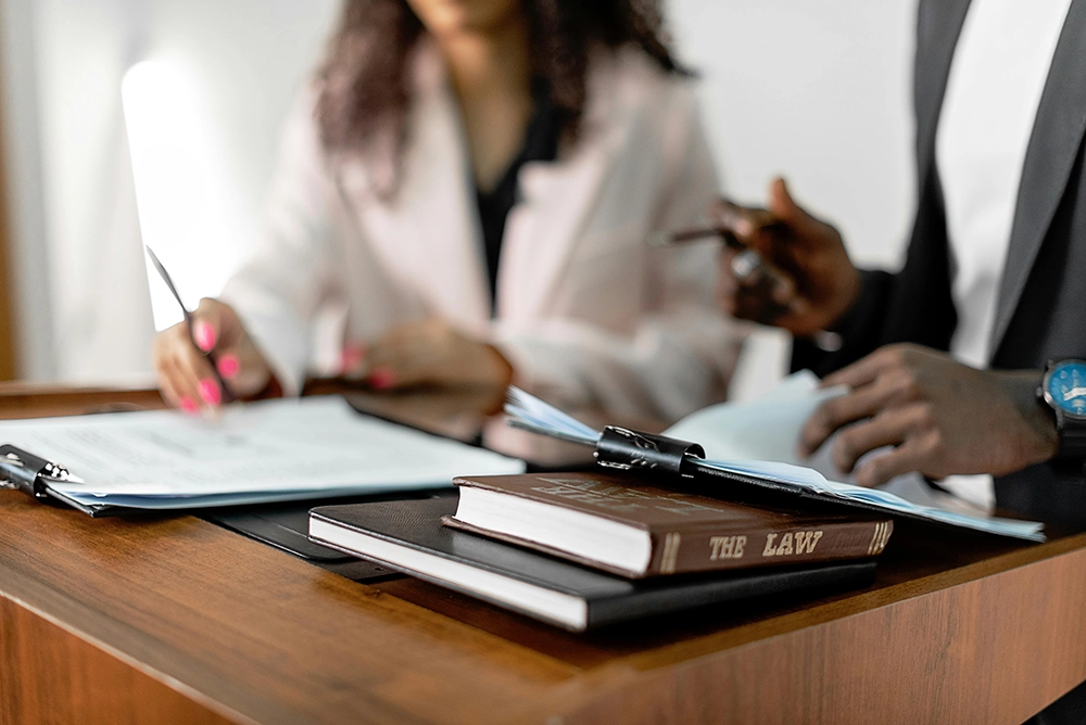 Two people review documents at a desk with a book titled "The Law" and papers in the foreground.