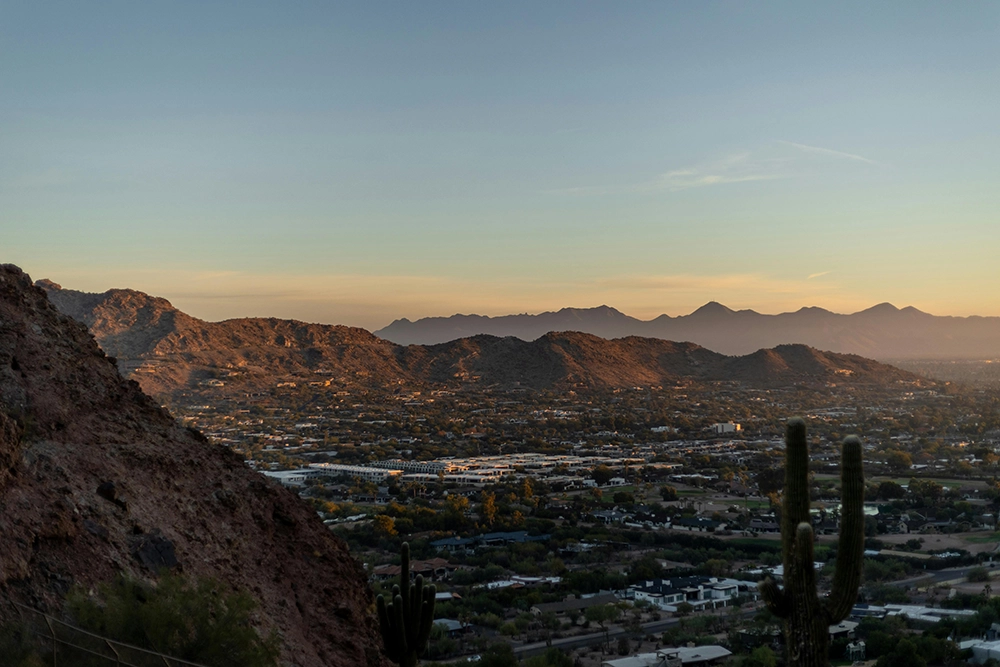 A view of a desert cityscape at sunset with mountains and cacti in the foreground and low buildings spread across the valley.
