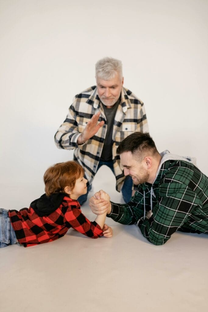 A child and an adult arm wrestle on the floor while an older man, raising his hand, looks on. All three wear plaid shirts against a plain background.