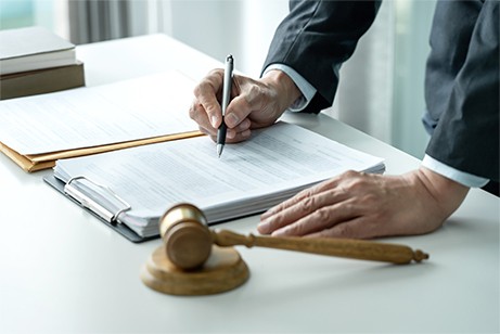 Person in a suit signs a document on a clipboard next to a wooden gavel on a desk.