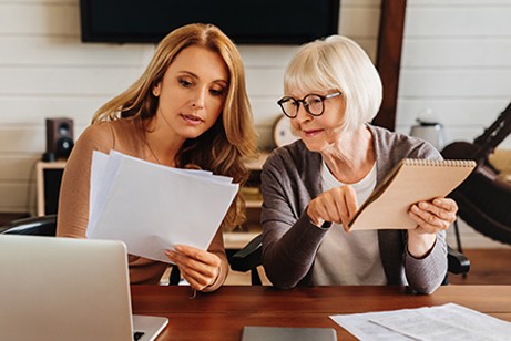 Two women sitting at a table, reviewing documents together; one holds papers while the other points to a notepad. A laptop and paperwork are on the table.