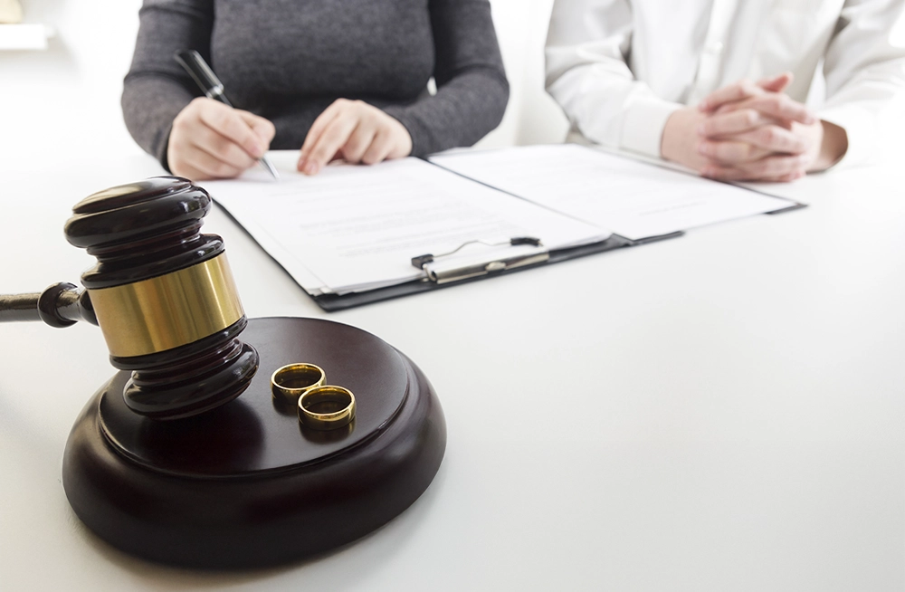 A gavel and two wedding rings on a table in the foreground, with two people signing divorce documents in the background.