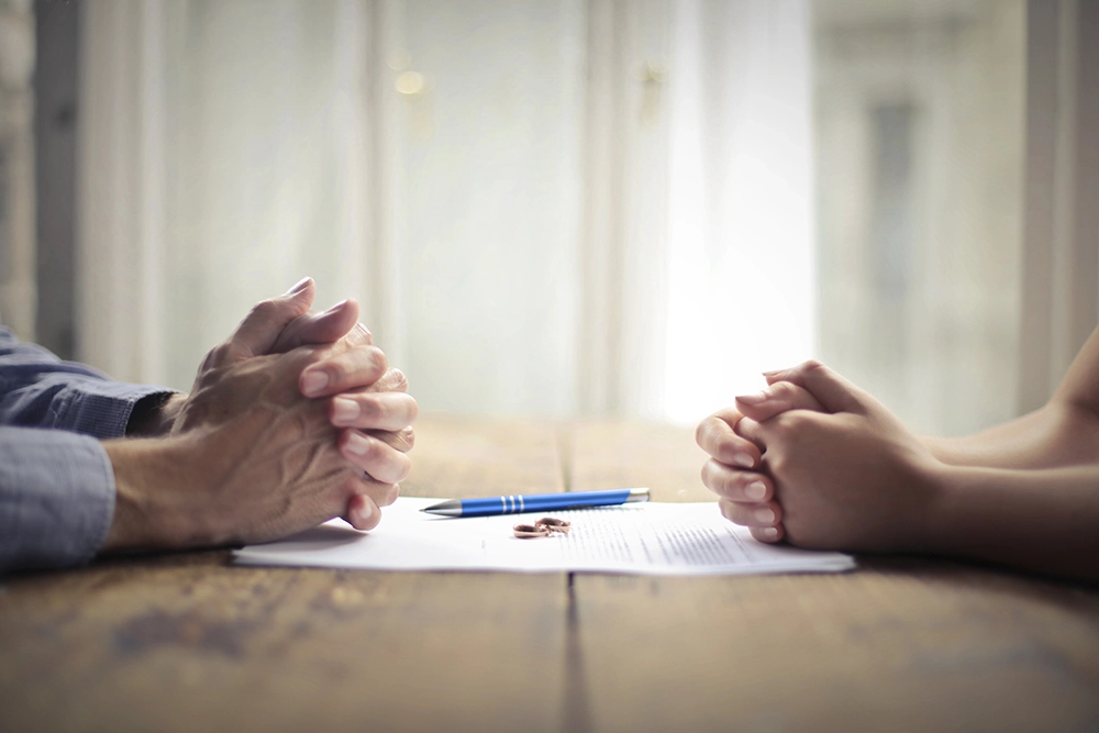 Two people sit across from each other at a wooden table with hands clasped, a document, pen, and rings placed between them.