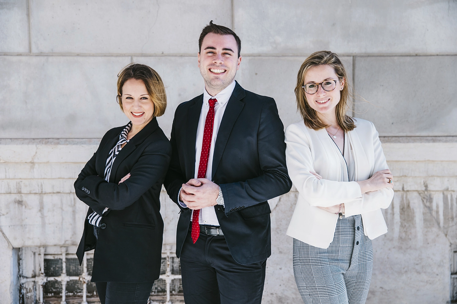 Three professionally dressed people stand side by side in front of a light-colored wall, smiling and facing the camera.