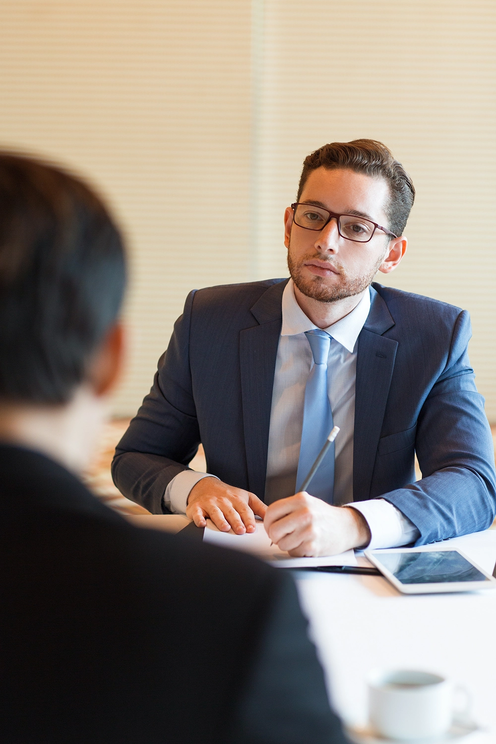 A man in a suit with glasses sits at a desk, holding a pen and notepad, listening to another person during a meeting or interview.