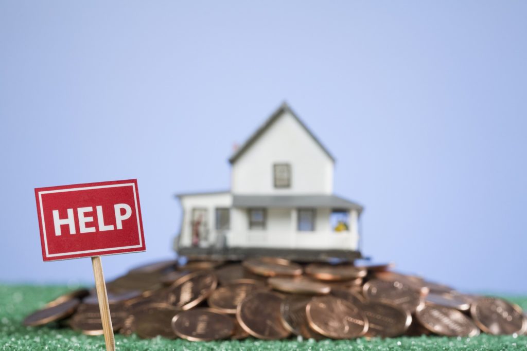 A small model house sits on a pile of coins with a red "HELP" sign in the foreground, suggesting financial distress related to housing—an issue where consulting a Gilbert Trust Attorney could provide guidance.