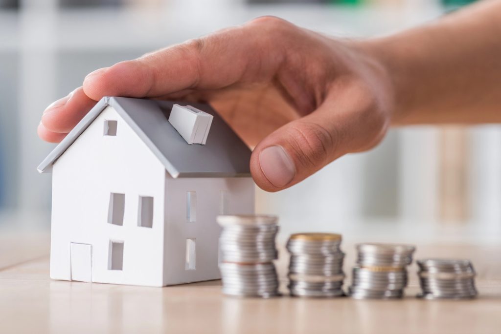 A hand covers a small model house next to four stacks of coins, symbolizing savings or investment in real estate—ideal imagery for a Chandler Estate Planning Attorney.