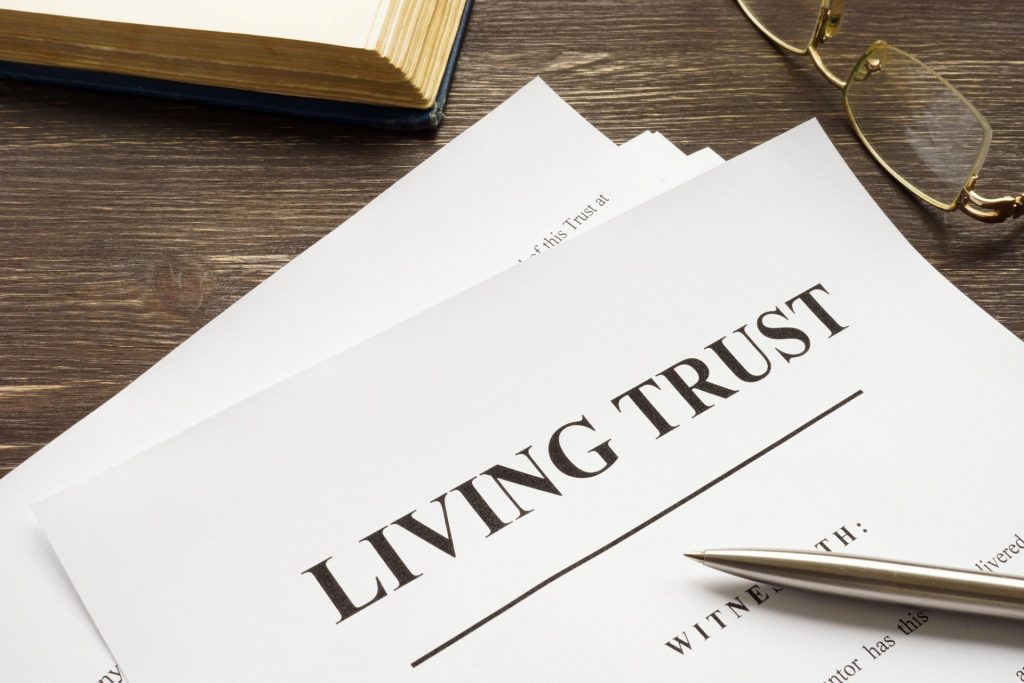 A stack of papers labeled "Living Trust" rests on a wooden desk with a pen, eyeglasses, and an open book nearby—ideal for discussing options with a Mesa Probate Attorney.