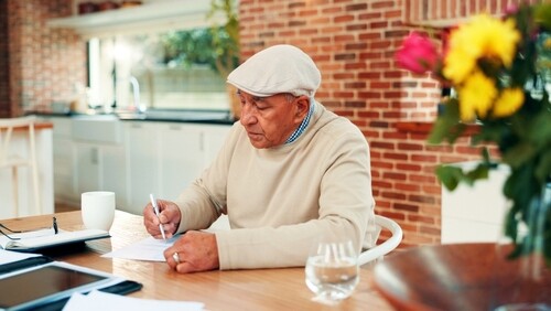 elderly man wearing flat cap writing on documents at home table