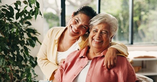 younger woman embracing smiling senior woman indoors near large window